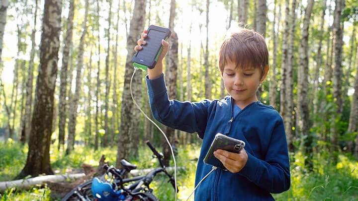 Ragazzo giovane con una giacca blu con zip su una pista ciclabile, con una bicicletta sullo sfondo, che solleva un piccolo dispositivo a pannello solare collegato a un telefono.