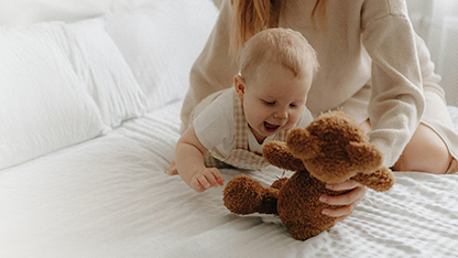 Immagine di una mamma che gioca con il suo bambino piccolo ed un orsetto di peluche, su un letto bianco